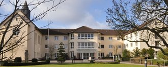 St. Josefshaus Über eine Rasenfläche blickt man auf ein U-Förmiges Gebäude in Beige. Auf dem Dach des linken U-Schenkels befindet sich ein Türmchen mit Kreuz. Mittig angeordnet ist der Eingangsbereich über dem der Schriftzug "St. Josefshaus" zu lesen ist.