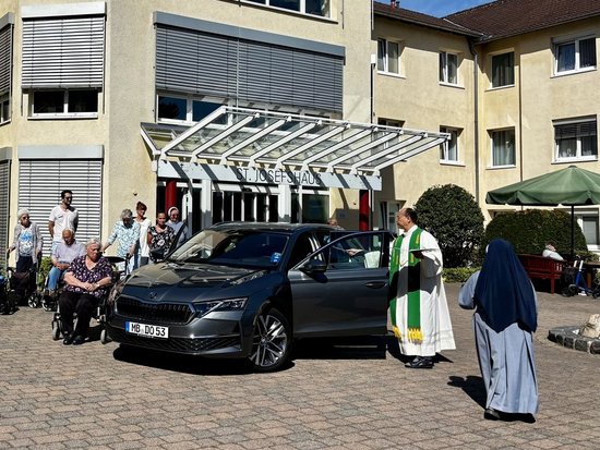 Ein Priester und eine Nonne segnen ein dunkles Auto auf dem Hof eines Pflegeheims. Ältere Menschen, teilweise im Rollstuhl, und weitere Personen beobachten die Zeremonie vor dem Gebäude.