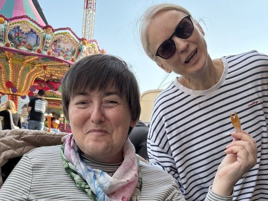 Zwei Frauen posieren auf einem Jahrmarkt. Eine Frau im Rollstuhl hält eine Schale mit Pommes und Nuggets. Eine zweite Frau mit Sonnenbrille steht lächelnd daneben und hält eine Pommes in die Höhe. Im Hintergrund ist ein farbenfrohes Karussell erkennbar.