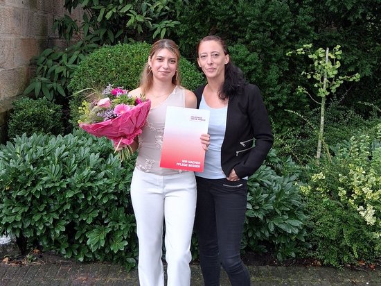 Zwei junge Frauen stehen vor einer Steinmauer und Hecken. Die linke Frau hält einen Blumenstrauß und ein Zertifikat, was auf eine Verleihung hindeutet. Beide lächeln in die Kamera.