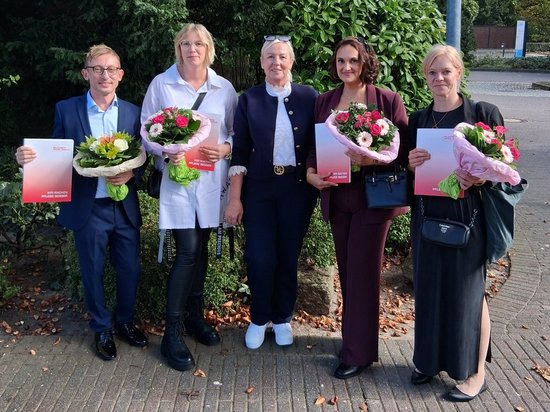 Fünf Personen stehen im Freien nebeneinander und halten Blumensträuße sowie rote Ordner. Es handelt sich vermutlich um eine Ehrung oder Preisverleihung. Eine Person trägt einen weißen Kittel, die anderen formelle Kleidung.